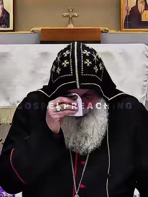 A close-up shot of a man, likely a religious figure, dressed in black robes and a dark, ornate hood. He is holding a tissue to his face, appearing to be wiping away tears. The background includes a cross and framed pictures, suggesting a religious setting.