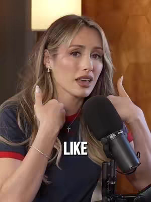 A woman with blonde hair and a navy blue top, wearing a gold cross necklace and a bracelet, smiles and gives a thumbs-up gesture with both hands. The text 'LIKE' is overlaid in white blocks. The background shows part of a lamp and a wooden surface, suggesting a podcast setting.