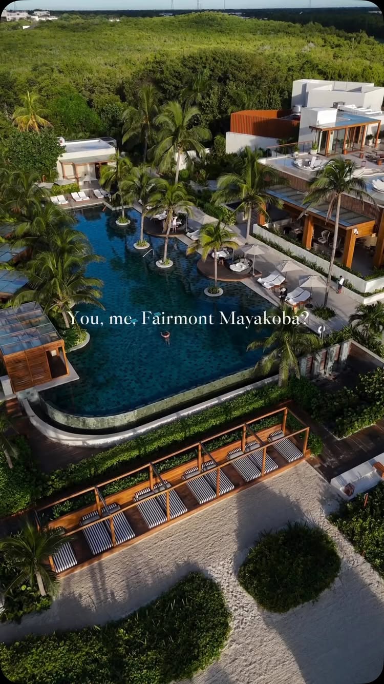 An aerial shot of a luxury resort pool area with a long, irregularly shaped pool surrounded by palm trees, buildings, and lush greenery. The focus is on the pool and its clear, dark water, hinting at a tropical location.