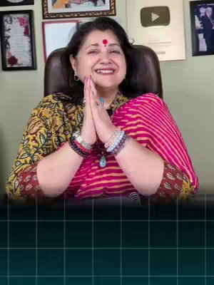 A woman in a brightly colored top smiles, with her hands in a prayer position. She has a red bindi and jewelry on her wrists. Behind her are framed pictures and a logo.