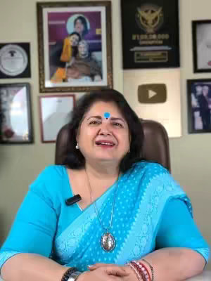 A woman is seated in a chair, facing the camera with a pleasant expression. She has dark hair, fair skin, and is wearing a light blue sari. Various framed pictures and awards are visible on the wall behind her.