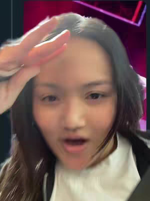A selfie-style shot featuring a young woman with long, dark hair. She is making a peace sign with her right hand, holding it up near her head. The open mouth suggest the creator is speaking or reacting to something. The background seems to be a dark, possibly indoor setting. The overall composition is close up.