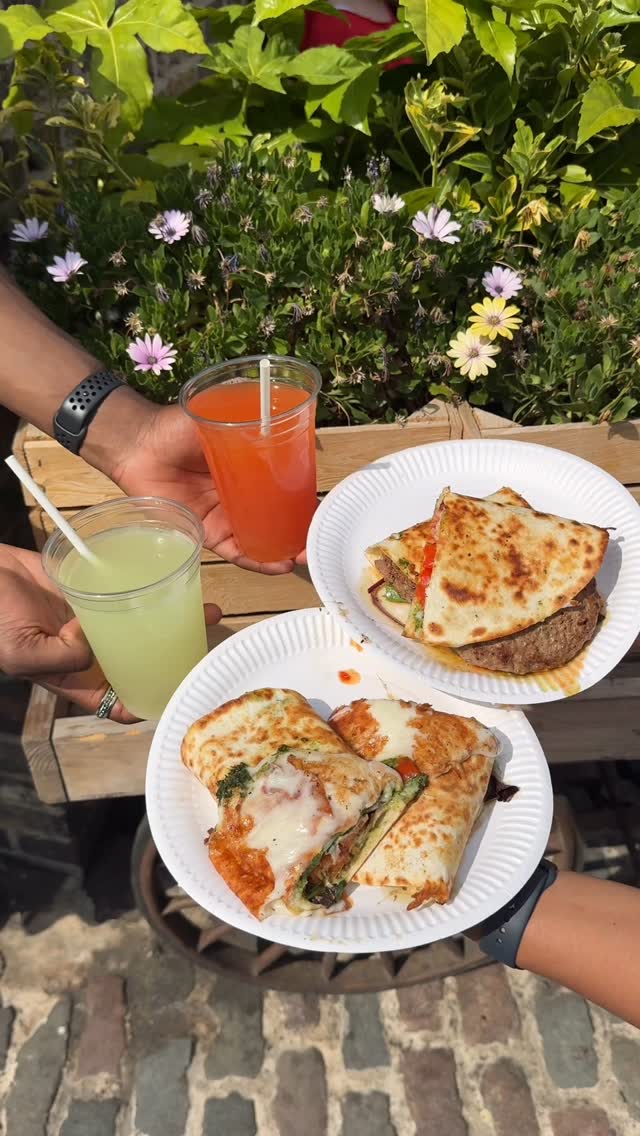 The image shows a close-up of three paper plates holding food items and drinks. Two plates hold folded flatbreads overflowing with fillings. There are also two clear plastic cups with colorful drinks, one yellow and the other a reddish-orange, both with straws. Greenery and flowers are visible in the background, giving a bright, outdoor appearance. The image is brightly lit and well-composed.