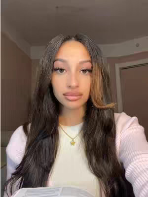 A woman with long brown hair, styled with layers, and asymmetrical highlights, poses for a selfie. She is wearing a white top, and a delicate gold necklace. The background seems to be a room with warm lighting.