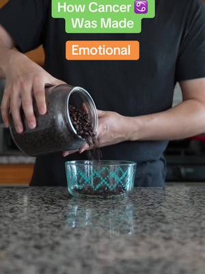 A person is shown from the torso up, pouring dark beans from a glass jar into a clear bowl. The person is wearing a black t-shirt, and the background includes a countertop and a kitchen setting. Text overlays at the top say "How Cancer Was Made" followed by the Cancer zodiac symbol