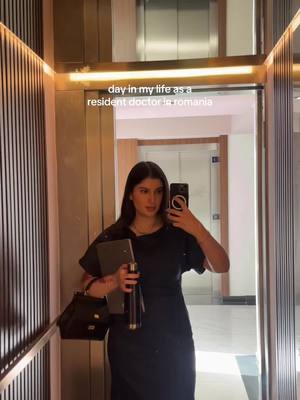 A woman stands in an elevator, holding a laptop and a water bottle with a handbag on her arm, taking a selfie with her phone, and looking in the mirror. There is text overlay on the top that reads "day in my life as a resident doctor in romania". The elevator surroundings are made of reflective materials with metallic details behind the woman within the elevator. The color palette is muted with tones of gold, gray, black, and neutrals, with the woman wearing a black dress. The layout is a straightforward, symmetrical composition showing the subject in the center.