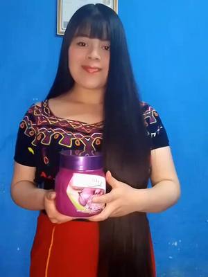 A woman with long black hair is standing in front of a blue wall. She is wearing a patterned top with a red skirt and is holding a jar of purple cream. She has a smiling expression.