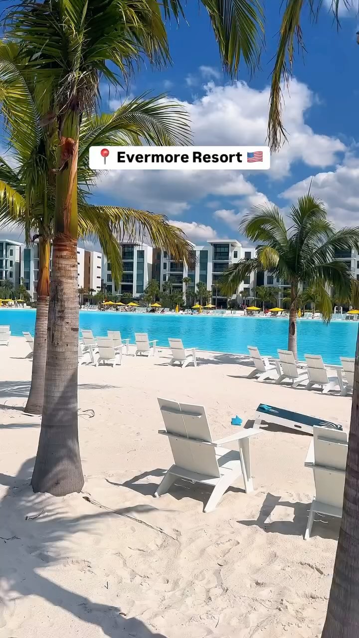 The image showcases a sunny resort scene at Evermore Resort. A large turquoise pool dominates the center, surrounded by a white sandy beach and lined with white lounge chairs. Palm trees frame the scene, and modern white buildings with glass panels are in the background. The sky is blue with scattered white clouds.