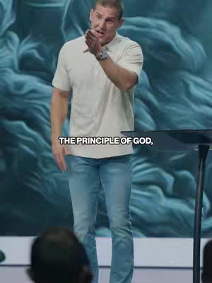 A man in a white button-down shirt and jeans stands behind a lectern, gesturing with one hand. The background features a blue, swirling design.