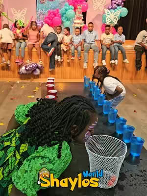 A group of children are playing a party game. The floor shows a table with red cups and a basketball net. Some kids are playing the game. In the background, other kids and adults are watching, which include many colorful balloons and celebration decorations