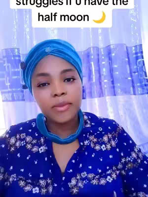 A woman with brown skin is positioned in front of a pale blue curtain in a selfie-style shot. She's wearing a vibrant blue headscarf and a matching top with a floral pattern in white and blue. The background is a light blue curtain providing a simple backdrop.