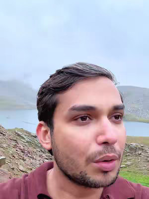 A man is looking slightly to his right and slightly up in the direction of the camera. The background features a mountain range with what may be a lake, all under an overcast sky.