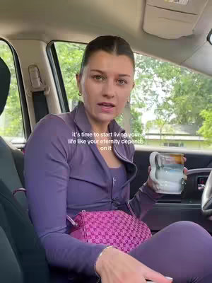 A woman in a purple tracksuit sits in the driver's seat of a car, holding a mug. The background shows a blurred street scene. Text overlay says 'it's time to start living your life like your eternity depends on it'.