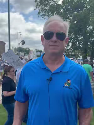A man with sunglasses and a blue polo shirt stands outdoors with a crowd in the background. Trees and a cloudy sky provide the backdrop.