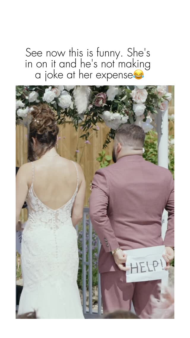 A wedding scene where the bride is in front looking away while the groom, with his back to the camera, is holding a sign that says 'HELP!' behind her back.