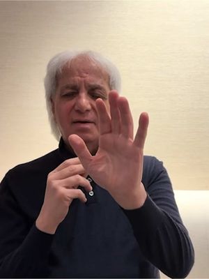A man with white hair extends his right arm toward the camera, covering his face with his hand. He wears a dark blue button-down shirt. The background is a simple, light-colored wall.