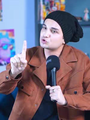 A man with short hair is pointing with his index finger while holding a microphone. He is wearing a black beanie, brown suede jacket, and a dark t-shirt. The background fades from light blue to a lighter color.