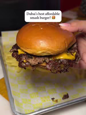 A close-up shot of a smash burger held by a hand. The burger has a toasted bun, a patty with melted cheese, and is placed on a paper liner within a metal tray. A white, rectangular text box in the top center reads "Dubai's best affordable smash burger?" The background is slightly blurred, suggesting a restaurant setting.