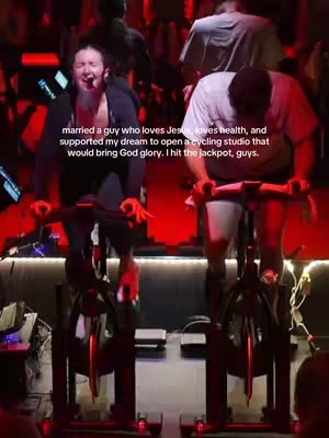 The image shows two people, presumably a couple, wearing activewear and riding stationary bikes in what appears to be a cycling studio. A person on the left is facing the camera with an open mouth, appearing to be shouting or singing, with the person on the right focused on cycling. Both are sweating and appear to be working hard. The background is dimly lit with red ambient lighting.