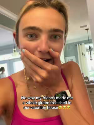 A selfie-style video cover image featuring a young woman with fair skin, wide blue eyes, and blonde hair pulled back. She wears a hot pink tank top and a gold necklace. Her mouth is open in surprise, and she is holding her hand over her mouth, covering it. Text below reads, 'No way my friends made me a whole gluten free shelf in our vacation house'.