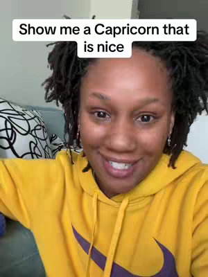 A woman with dark hair and a yellow hoodie smiles towards the viewer. Text overlayed at the top reads 'Show me a Capricorn that is nice.' The background is blurred, featuring a portion of a patterned cushion.