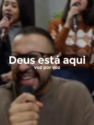 The cover image presents a blurred, close-up view of a man with glasses and a beard, holding a microphone. The text "Deus está aqui" (God is here) is prominently displayed in white, bold font. Beneath, in a smaller font, it says "voz por voz" (voice by voice). Other people with microphones and visible backgrounds are also present. The image has a slightly out-of-focus or soft focus effect, contributing to the impression of a background of people. The overall composition has a warm, inviting feel, centered on the man in the foreground.