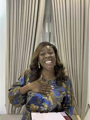 A woman with long brown hair and holding her hand to her chest is smiling in front of a window with vertical cream-colored curtains. She's wearing a patterned top, and has gold earrings. She is holding a tablet, and is lit with soft natural light.