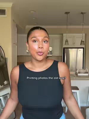 A woman is standing in a kitchen setting, facing the camera and slightly pouting, with a text overlay reading "Printing photos the old way?". The scene includes a bright kitchen with white cabinetry.