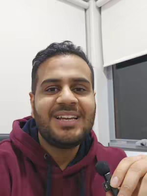 A close-up shot of a man smiling directly at the camera. He has short, dark hair and a beard. He's wearing a dark red hoodie over a dark blue shirt. In his right hand, he holds a small microphone.