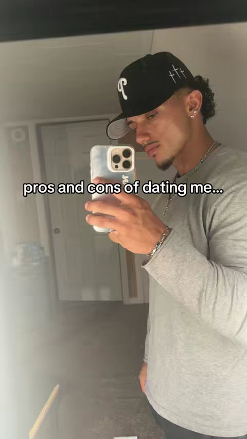 A man is seen against a mirrored background, holding a phone and looking towards the camera with a composed expression. He is wearing a grey long-sleeved shirt and a black cap, with the text 'pros and cons of dating me...' overlaid.