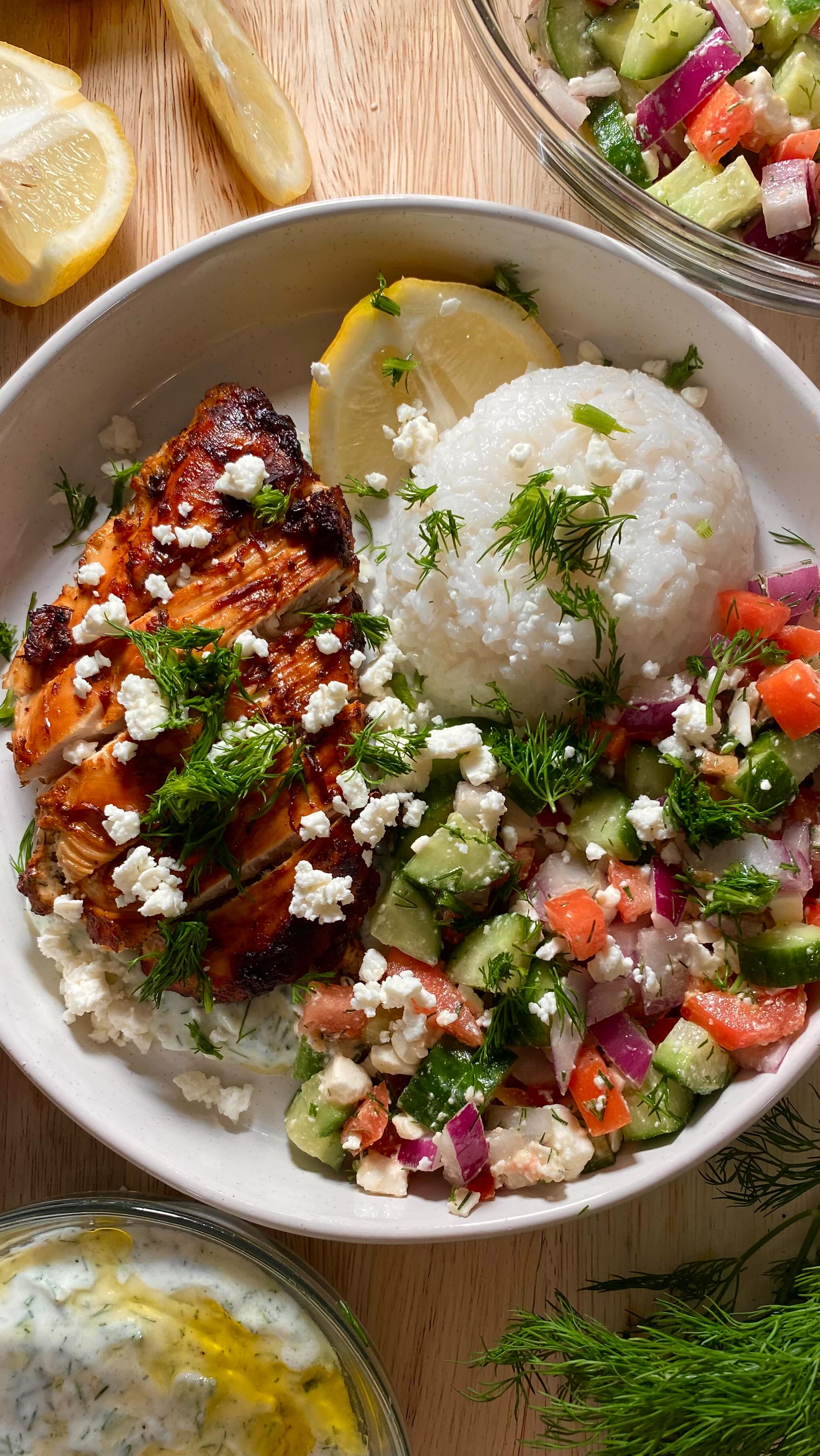 A top-down shot of a plate of food. The plate is filled with chicken, rice, and a salad. A lemon wedge is nestled on the side. The food is arranged on a white plate set against a wooden surface, creating a warm, inviting presentation.