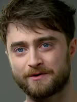 A close-up shot of actor Daniel Radcliffe against a gray background. He has short, dark hair styled to the side, blue eyes, and a full beard. He appears to be speaking, with his mouth slightly open. The lighting is soft and even, and the focus on his face creates an intimate feel.