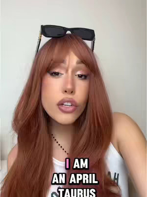 The cover image features a woman with auburn hair and styled bangs, wearing a white top. She has sunglasses perched on her head, and text overlayed on the image reads "I AM AN APRIL TAURUS". The background is a plain white wall. The woman is looking at the camera with a slight pout and her lips done with nude lipstick and perfectly made-up eye.
