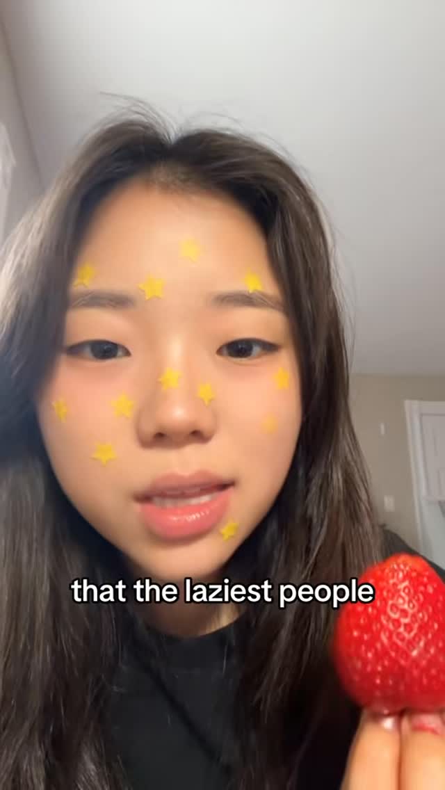 A young Asian woman is featured in a selfie-style shot. She holds a red strawberry in her hand, with the fruit taking up the lower right portion of the frame. Her face has multiple star-shaped stickers scattered on her cheeks. The text overlay at the bottom of the frame says 'that the laziest people'.