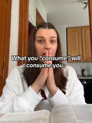 A person is visible from the chest up, with light brown hair. The person, who appears to be the creator, is wearing a white sweatshirt and has their hands clasped in front of their face. The shot is centered, with a blurred book in the foreground. Behind the person, there's a wooden frame, and some cabinets. The text overlay states, 'What you “consume” will consume you..' .