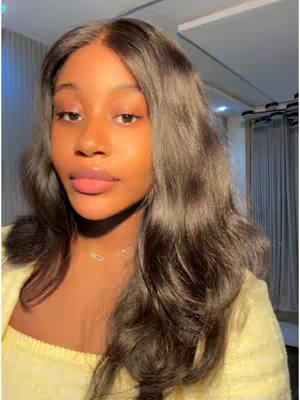 A close-up shot of a young woman with dark wavy hair. She's wearing a yellow top and a delicate necklace. The background is slightly blurred, revealing a room with curtains and a hint of indirect sunlight.