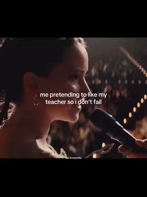The image is a still from a scene, featuring a woman speaking into a microphone. She has dark hair with a side part and is wearing earrings. She is positioned in profile with a focused expression, suggesting she may be on stage. The lighting is dim, further emphasizing the woman's face. The background is out of focus but appears to be a large auditorium or stage.