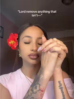 A woman with dark hair and a red flower in her hair is in frame, her eyes are closed, and her hands are clasped in a prayer position. Text overlay at the top says, Lord remove anything that isn't-