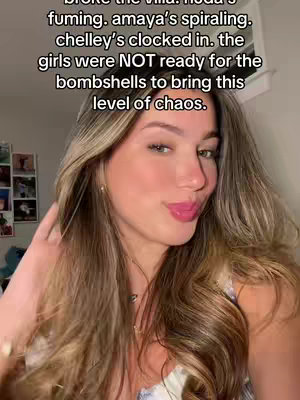 A selfie-style shot featuring a young woman with long, wavy blonde hair. She is looking at the camera with a slight smile, her lips are glossy and pink, and she is wearing a light-colored top. In the background, there is a hint of a wall with some pictures, an overhead light, which suggests an interior setting.