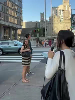 A woman is speaking into a microphone on a city street with two people standing nearby, and people walking in the background. The woman is the focus, holding a mic. A background shows a crossroads with cars, buildings, and pedestrians, with a distinctive architectural style.