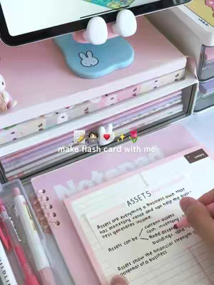 The image showcases a well-organized study setup with a pink aesthetic. A tablet is propped up on a bunny-shaped holder, which is positioned above a stack of notebooks and papers. A hand is writing on a note sheet, with a pen. The overall composition is clean and inviting, suggesting an environment conducive to studying. The scene is bright and well-lit