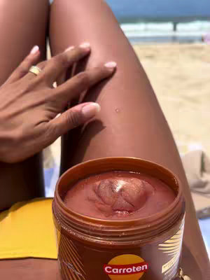 The image shows a close-up of a person's legs with one hand on the leg and a jar of tanning gel in the foreground. The legs are tanned with a slight shine from the product. The person's hand, with a ring, is lightly touching the skin. The jar on the ground reveals the product's contents, and a beach scene forms the backdrop.