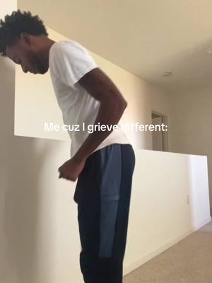 A man, wearing a white shirt and dark blue and black track pants, leans his head against a white wall in a hallway. Text overlay at the bottom reads "Me cuz I grieve different:". The hallway has tan walls and a light colored floor. There is a door in the background and a banister on the opposite side.