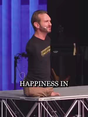 The image features a man with no arms and legs, centered on a stage, speaking with a microphone. Text overlayed on the image says "HAPPINESS IN". The background includes blue and purple lighting.