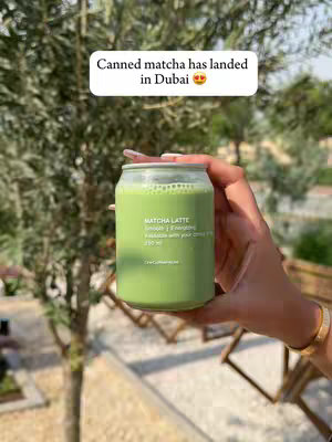 A hand holding a green matcha drink in a clear glass can. The background features blurred greenery and a hint of a wooden structure. The text overlay reads 'Canned matcha has landed in Dubai'.