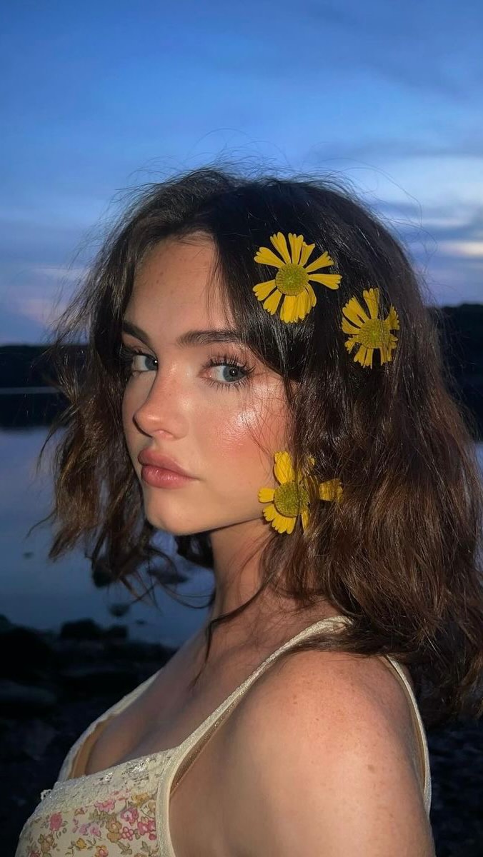 A close-up shot of a young woman with long brown hair, styled with large yellow flowers tucked behind her ear and in her hair. Her skin has a dewy glow, and she's wearing a light-colored camisole. The background is a blurred dusky blue, implying a twilight or nighttime setting.