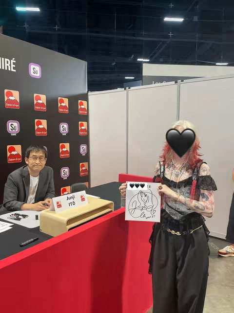 A person with red and blonde hair and tattoos in a complex outfit holds a drawing with a signature, standing in front of a booth with Junji Ito branding at Japan Expo. Behind the person is a man sitting at a table with a sign that says "Junji Ito". There are red walls with Japan Expo branding as well as other branding.