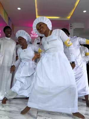 A group of people in white religious garments are dancing. The image is brightly lit, set indoors with a neon sign in the background. Several people are in focus toward the front, mid-movement, giving the impression of a lively performance.
