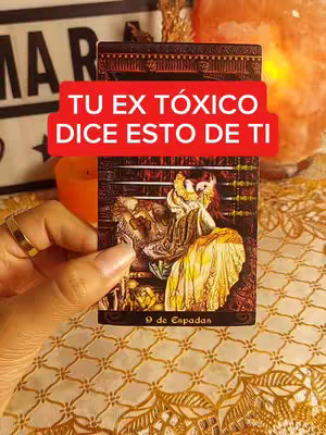 A hand holding a tarot card with an image of a woman in a long, elaborate dress. A red banner with white text 'TU EX TÓXICO DICE ESTO DE TI' (Your toxic ex says this about you) is at the top. The card is set against a blurry background of a candle, crystals, and fabric.