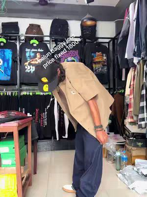 A person wearing a Carhartt cap and tan shirt leans into a store, with clothing racks and merchandise in the background. Text overlay mentions 'balun baggy jeats' and a price, with a focus on the casual aesthetic.